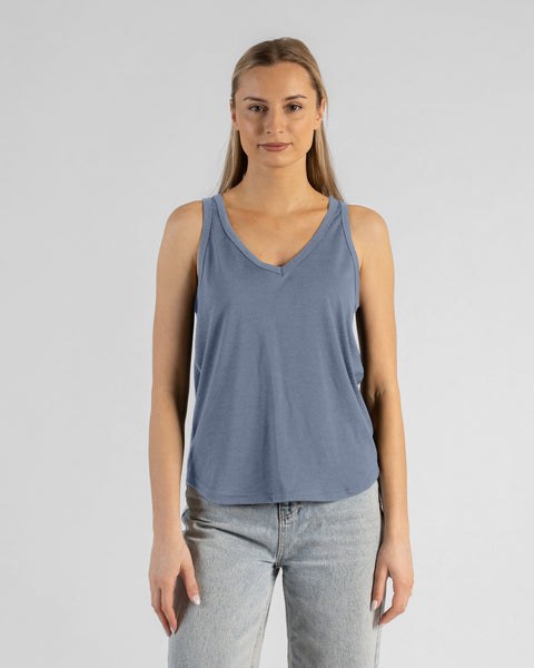 A studio portrait of a blonde woman standing and looking directly at the camera with her arms relaxed by her sides. She is wearing a regular fit, dusty blue sleeveless v-neck top and light-wash blue jeans against a plain light grey background.