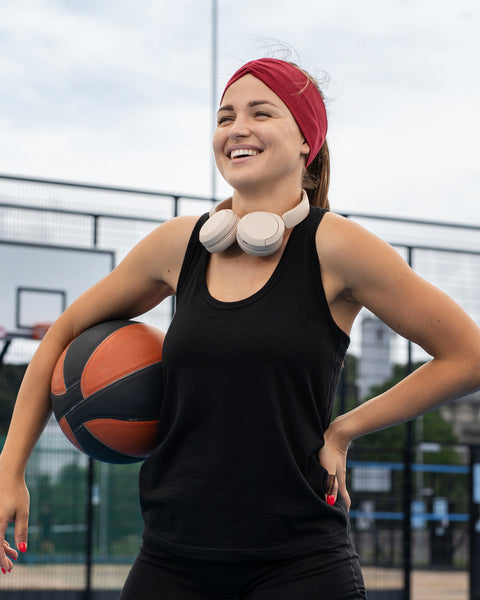 A smiling woman standing on an outdoor basketball court holding a basketball under her arm with her other hand on her hip, wearing a black tank top, a red headband, and white headphones around her neck.
