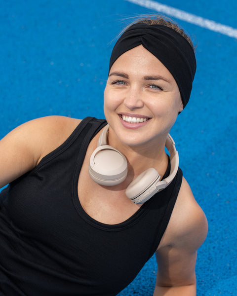A close-up of a smiling woman on a bright blue sports court, wearing a black tank top, a wide black headband, and white headphones resting around her neck.