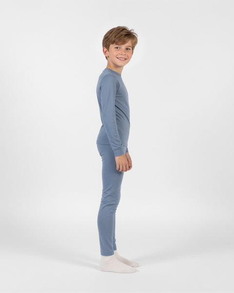 Young boy standing in profile looking toward the camera and smiling, wearing a dusty blue long-sleeve thermal base layer set and white socks against a plain white background.