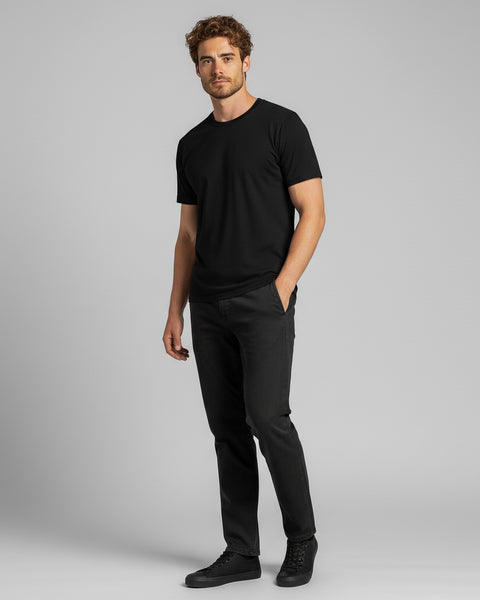 A man wearing a black T-shirt, black jeans, and black sneakers stands casually facing forward with one hand in his pocket against a light gray background.