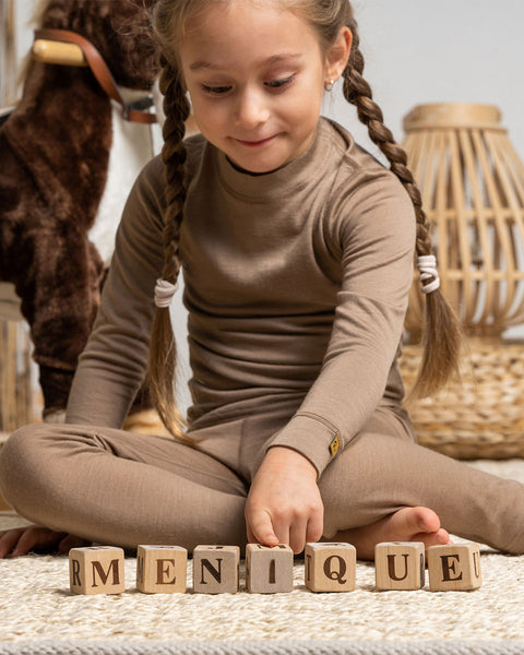 A girl, playing on the carpet with wooden cubes, wearing the menique Kids' Merino 160 Pants Beige color.