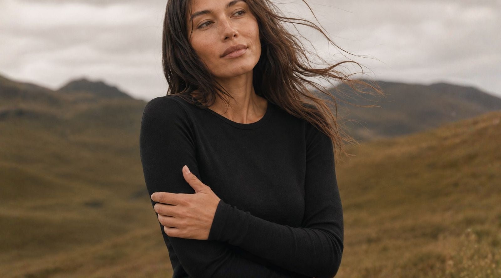 A woman wearing a black long-sleeved top standing in a windy mountainous area with her arms crossed.