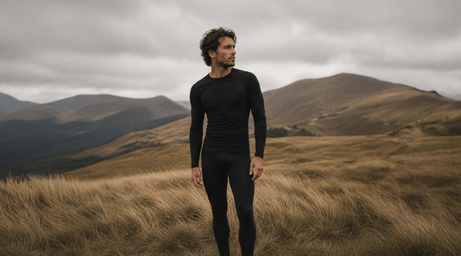 A man wearing a black long-sleeve compression shirt and matching black leggings stands on a grassy mountain ridge under an overcast sky.