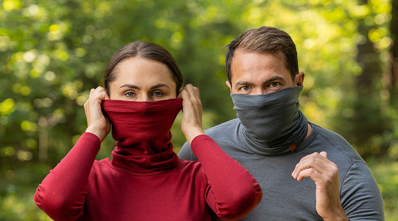 In this photo you can see man and woman standing in the forest. They are both wearing neck gaiters made from organic 100% Merino wool. Woman is wearing red color neck gaiter and Man is wearing grey one.