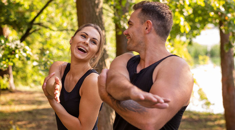 In this picture you can see man and a woman standing in the forest, streching and smiling at each other. They are both wearing black sleeveless tops made from natural 100% Merino wool.