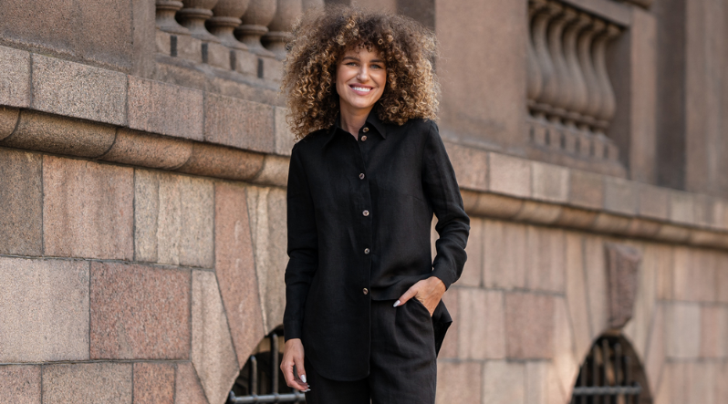 a woman standing by a building, wearing a full black linen set, that includes black pants and a black linen button-up shirt. She is smiling and has curly hair.