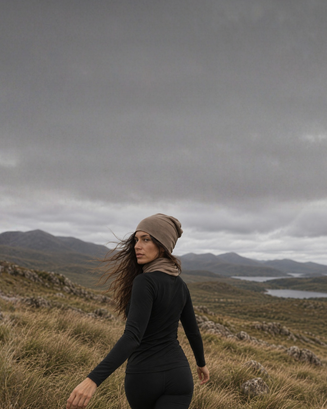 A woman with long wavy hair looks back over her shoulder while walking through a grassy mountain landscape. She is dressed in a black long-sleeved top and leggings, accessorized with a matching tan beanie and neck gaiter under a moody gray sky.