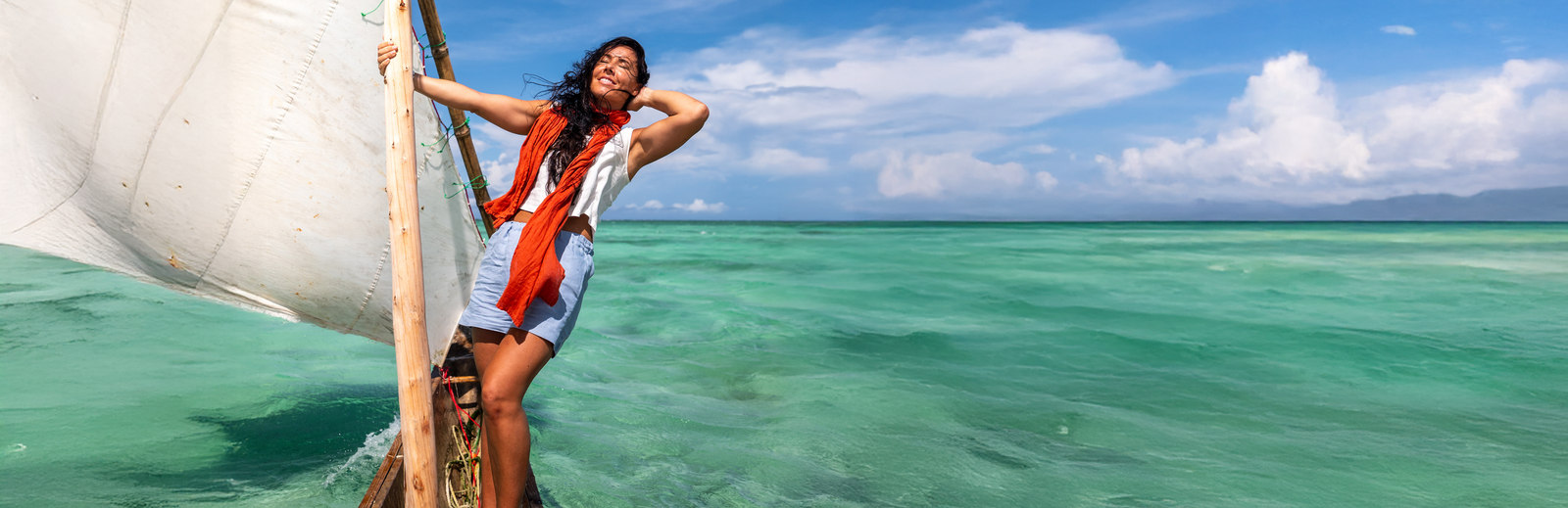 A woman standing on a traditional wooden boat, holding onto the mast with a white sail to her side. She is smiling with her eyes closed, enjoying the breeze, with one hand behind her head. She is wearing a white sleeveless top, light blue shorts, and a vibrant orange scarf blowing in the wind. Clear turquoise ocean water and a partly cloudy blue sky fill the background.
