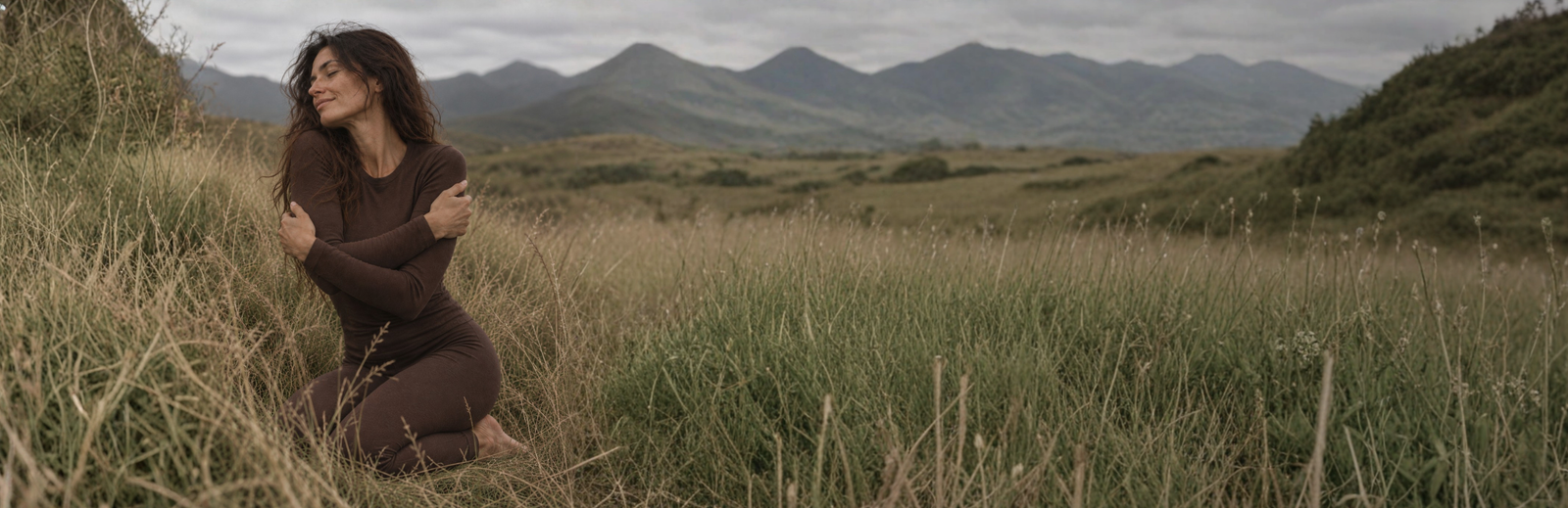 A woman kneels peacefully in a field of tall grass with her eyes closed and her arms crossed over her chest. She is wearing a brown long-sleeved thermal set, framed by distant mountains and a cloudy gray sky.