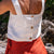 A woman with shoulder-length dark hair, wearing a pure white linen sleeveless crop top with button closures down the back and menique linen skirt, stands on a sandy beach looking over her left shoulder towards the ocean under a bright sky with scattered clouds.