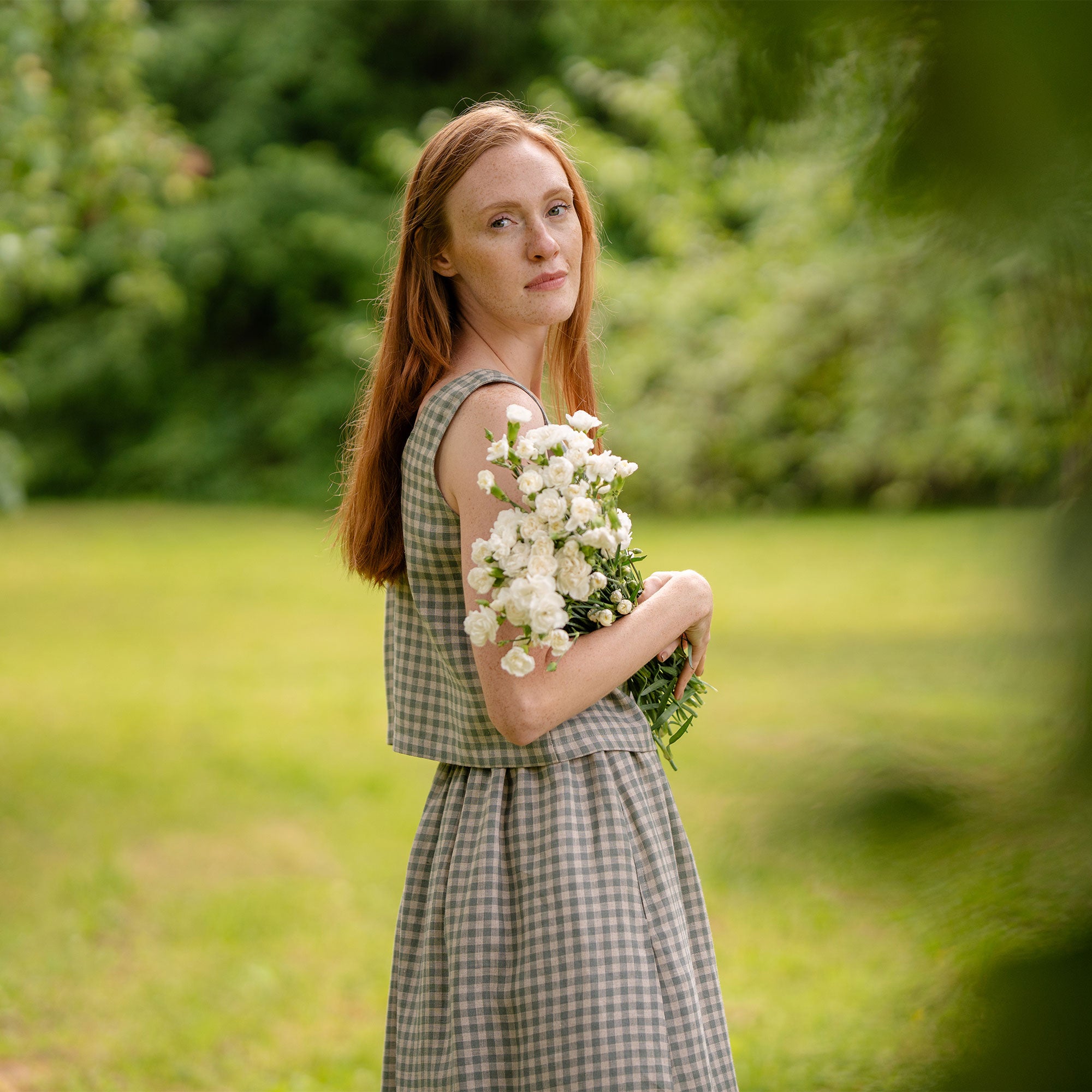 In this picture the model is standing in front of a clothesline with white garments hanging on it. Beside her is a metal bucket full of flowers. She is visible full-height, as well as the full skirt length. The skirt is knee length, matched with a same pattern blouse.