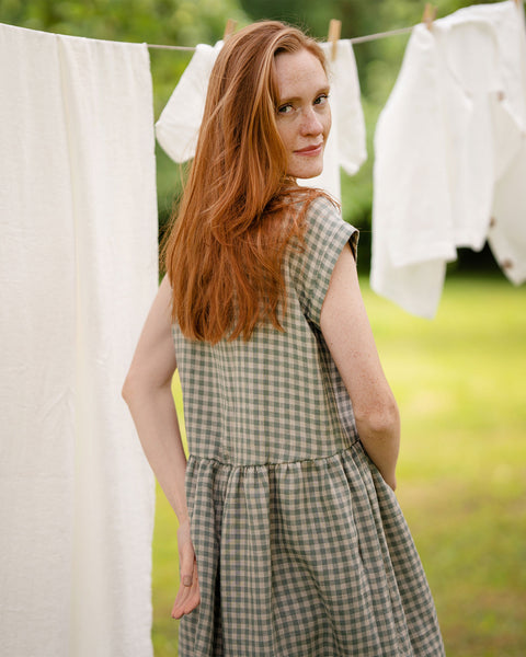 The model is wearing a light green and white gingham pattern dress, shown from the back in the photo. She is outside, there is laundry hanging behind her. The hem of the dress is dropped, asymmetrical. 