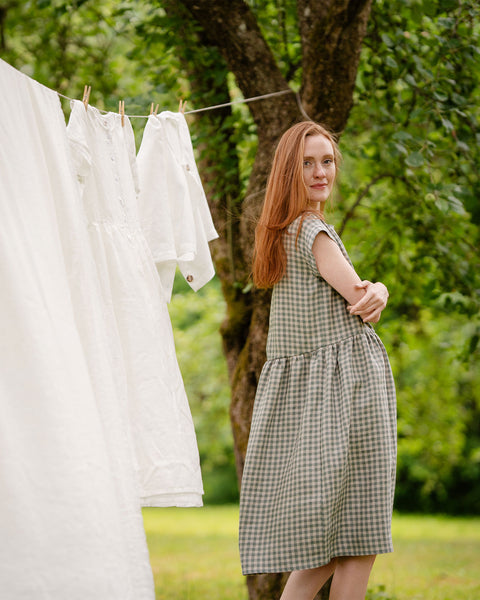The model is wearing a light green and white dress in a gingham pattern, standing sideways. She is smiling slightly with her palm put over opposite elbow. There are white garments hanging beside her.