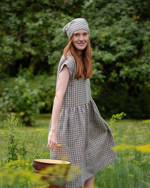 The model is smiling at the camera, standing sideways. She is holding a bucket in her hand in a field full of tall grass. She is wearing a green gingham dress and a matching bandana. 