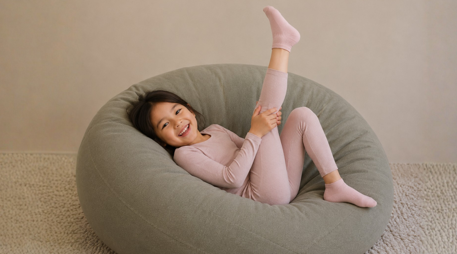 A young girl in a light pink long-sleeve pajama set and matching socks relaxes in a large, round, dusty green bean bag chair against a neutral beige wall.