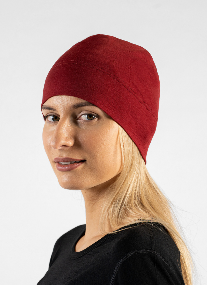 A blonde woman with a gentle smile looks towards the camera against a plain light grey studio background. She is wearing a snug, deep red knit beanie and a simple black crew-neck top.