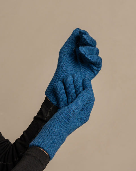 
A close-up shot of a person&#39;s hands wearing menique brand vibrant blue, ribbed knit gloves. The left hand is clenched into a fist, while the right hand appears to be gripping the wrist area of the left glove. The person is wearing a dark long-sleeved shirt underneath. The background is a solid, neutral gray.