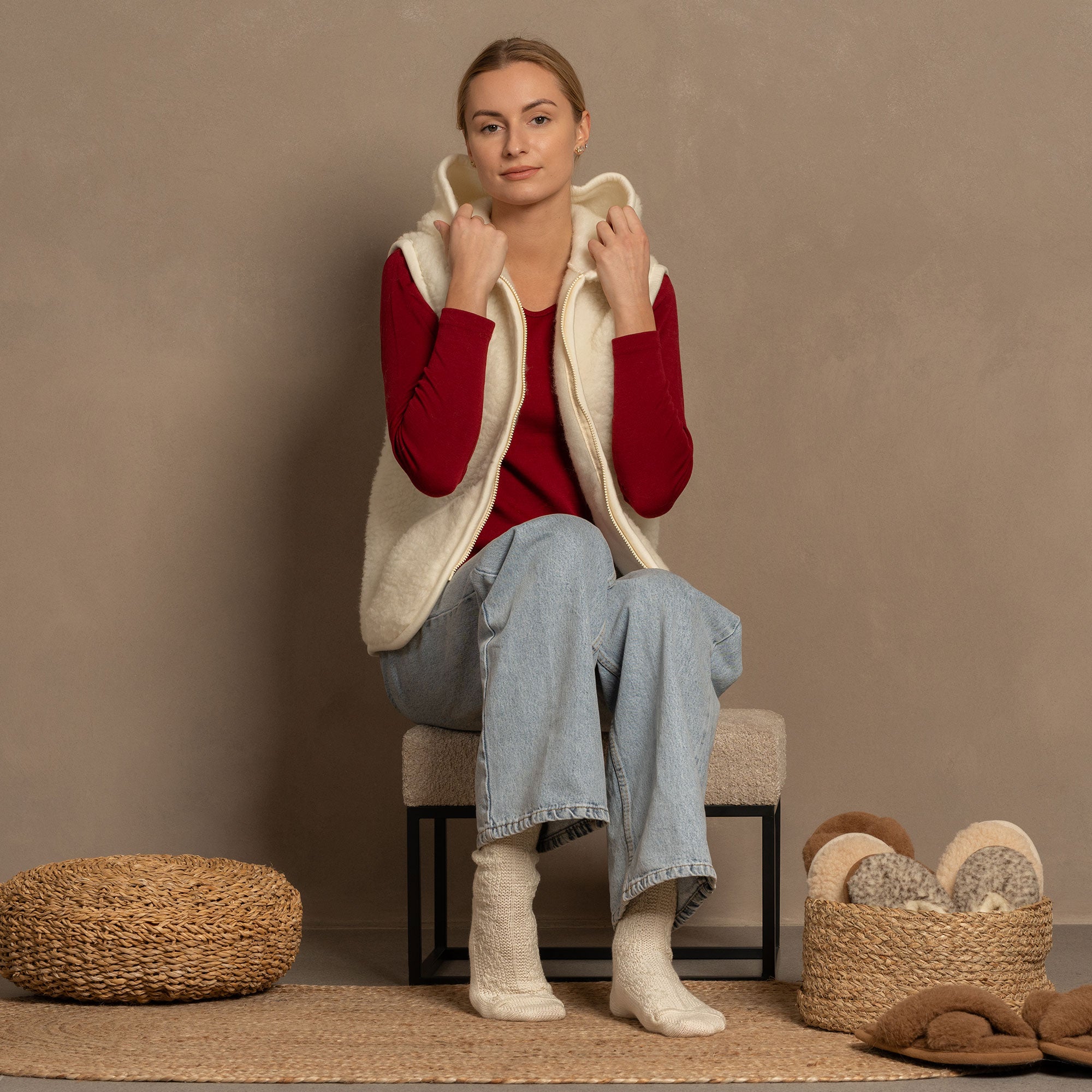 A low-angle shot shows a person's legs and feet, likely wearing menique brand off-white, crew-length knitted wool socks with a visible ribbed cuff and textured cable-knit pattern on the foot. The person is standing on a light brown, woven mat, against a neutral, plain background.