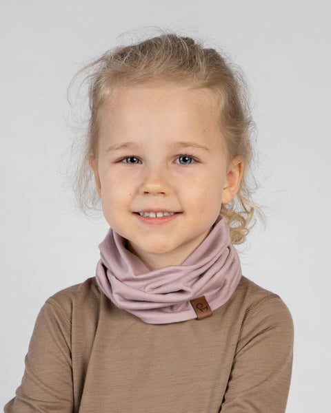 A close-up studio portrait of a young girl looking directly at the camera with a bright smile against a plain light grey background. She has blonde hair pulled back and is wearing a brown long-sleeved top with a dusty pink neck gaiter bunched around her neck, featuring a small brown logo tag.