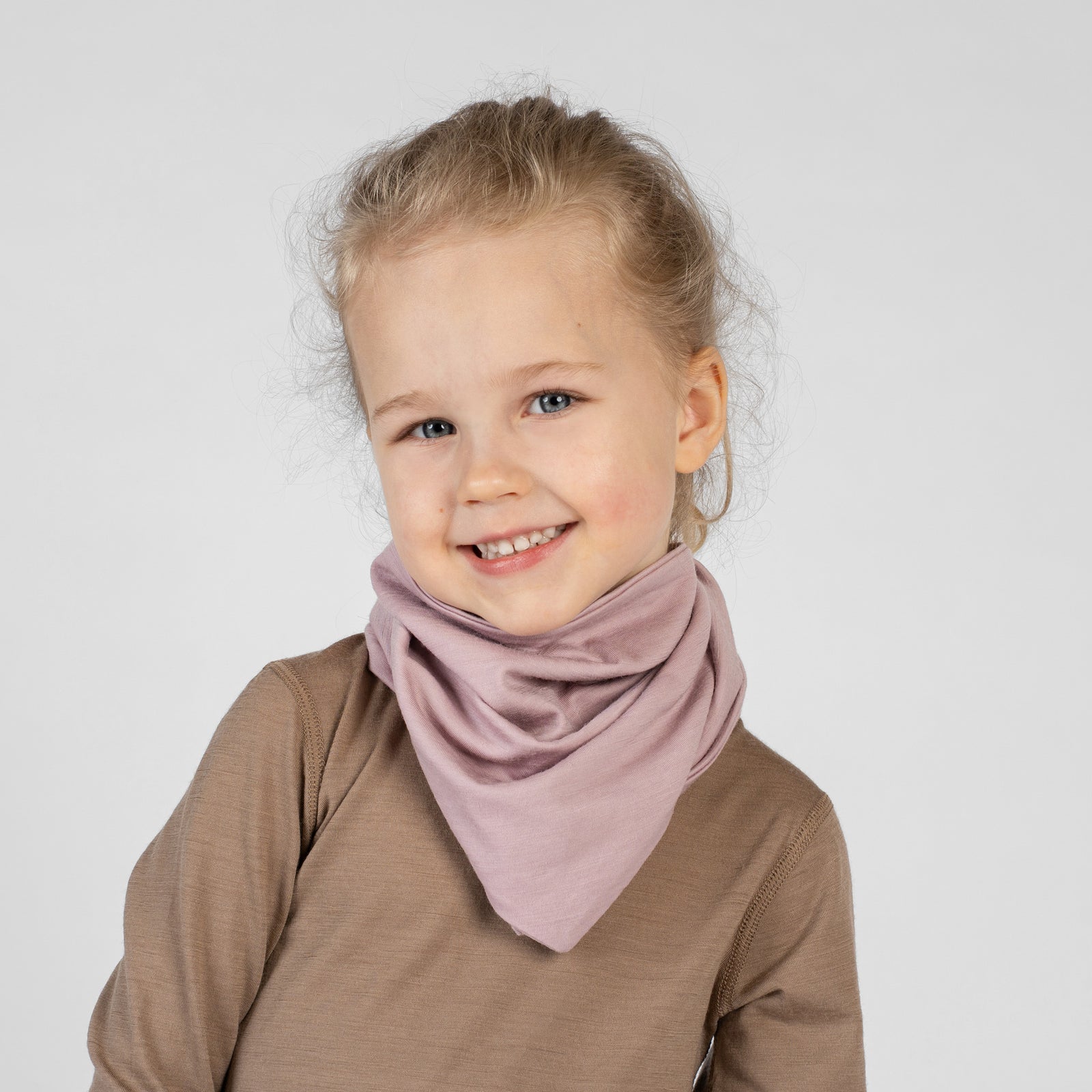 A studio portrait of a young blonde girl looking directly at the camera with a big smile. She is wearing a light beige bandana tied around her neck with the point facing down, over a solid black long-sleeved shirt. The background is a plain light grey.