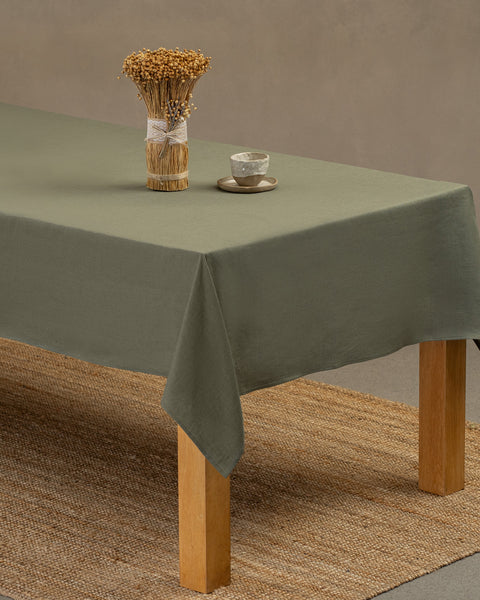 The menique stone green linen tablecloth on a wooden table. On top of the tablecloth is a vase made of natural straws and a clay cup. Under the table is a carpet.