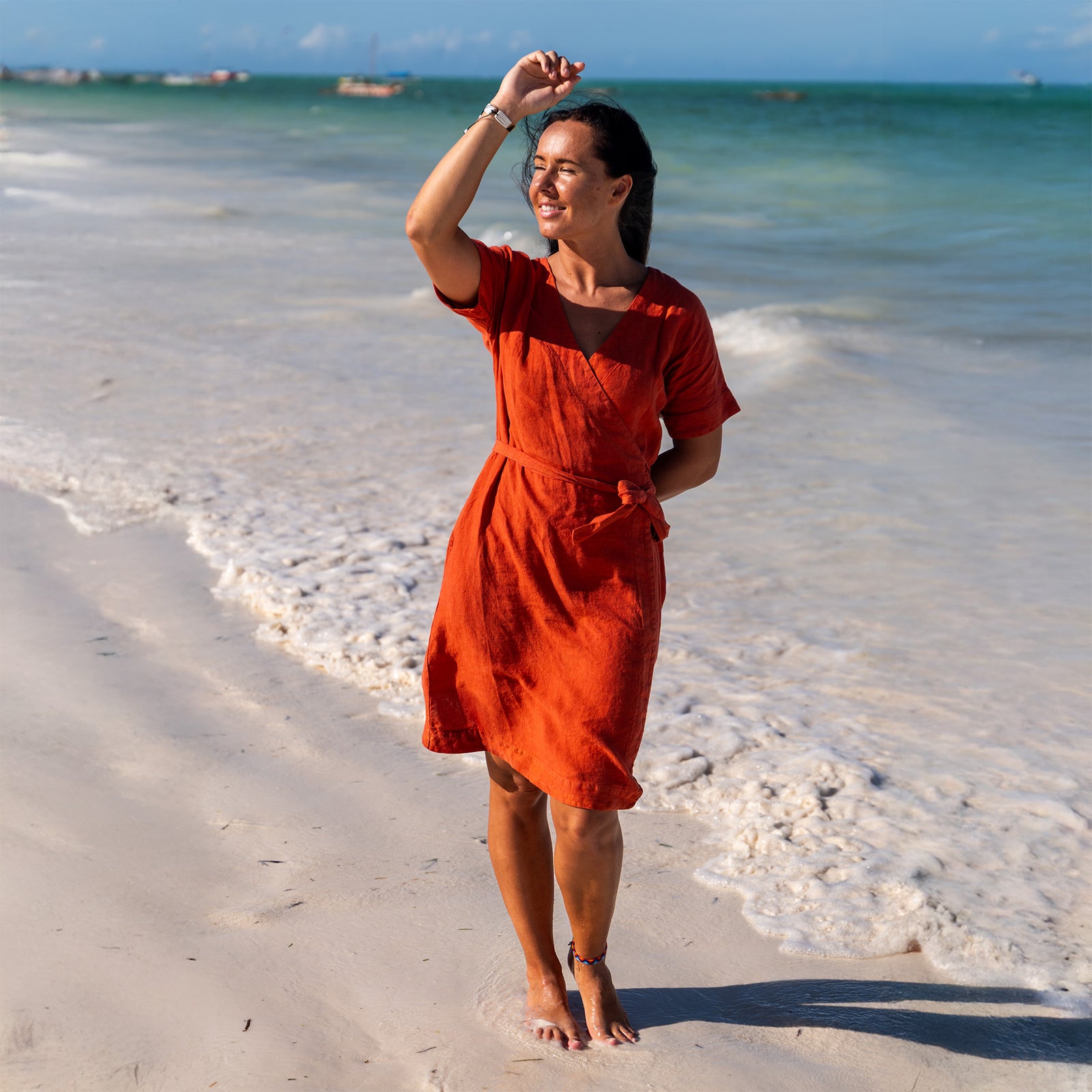 A smiling woman with long dark hair stands barefoot on a sandy beach, wearing a cinnamon nred linen sundress with thin straps. Her arms are outstretched to the sides. The ocean is in the background with gentle waves, and several boats are visible in the distance under a bright sky.
