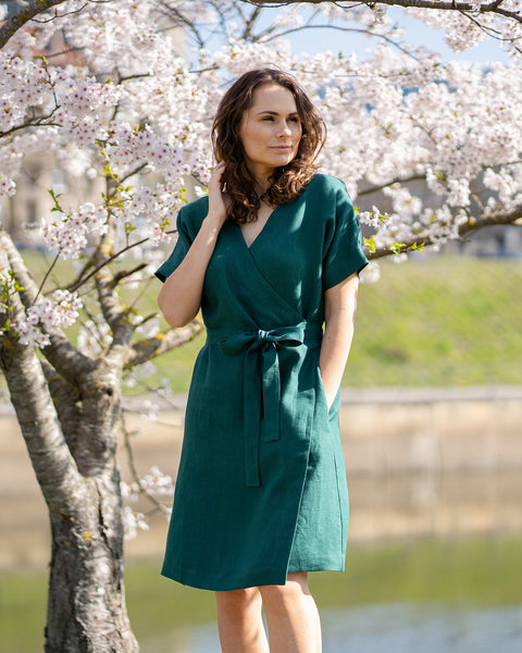 A woman with brown hair, wearing a dark green, short-sleeved wrap dress, stands outdoors under a tree with white blossoms.