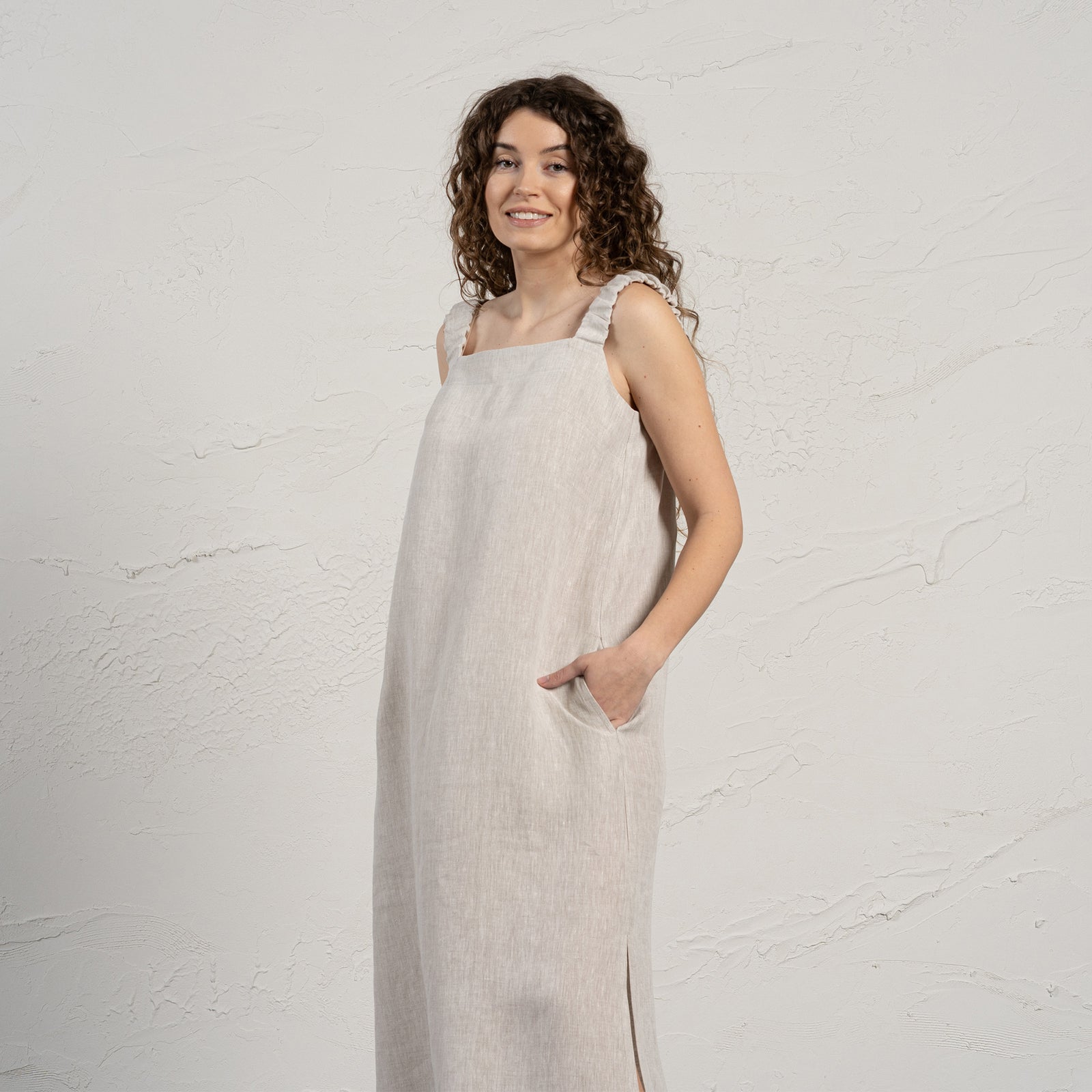 A light-skinned woman with long, dark curly hair stands facing the camera with a gentle smile. She is wearing a sleeveless, menique maxi dress with a square neckline and side slits, and black sandals. Her hands are in the dress pockets, and she stands on a light gray floor against a textured off-white wall.