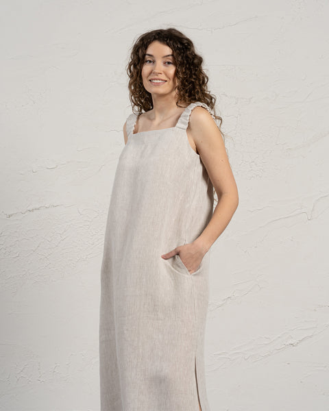 A woman with light skin and long, dark curly hair smiles gently while looking slightly to her right. She is wearing a sleeveless menique maxi dress with a square neckline, and her right hand is in the dress pocket. She stands against a textured off-white wall.