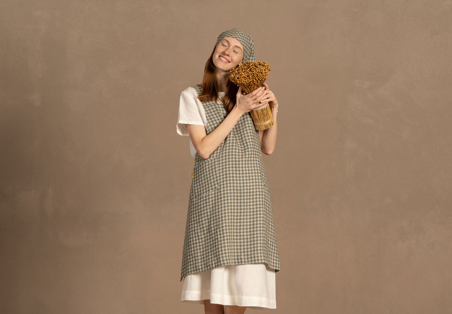 A smiling woman wearing a green and white checkered linen apron and matching headscarf over a white dress, hugging a bundle of dried flax.