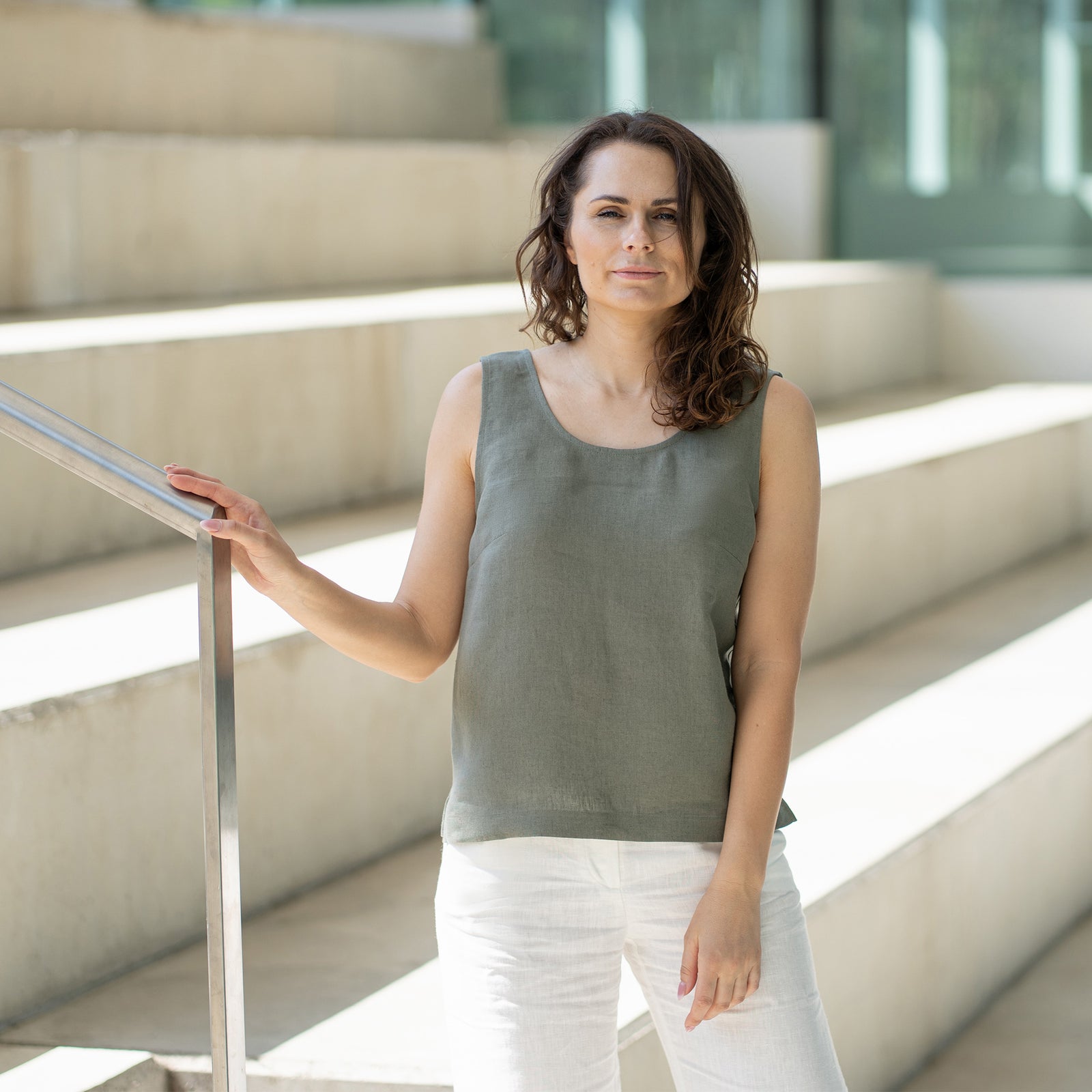 A woman with brown hair, wearing a stone green sleeveless linen top and white trousers, stands on wide concrete stairs inside a modern building with glass walls.