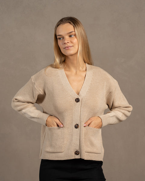 A woman, looking to the side, wearing a beige merino wool knit cardigan. She has both her hands in the front pockets.