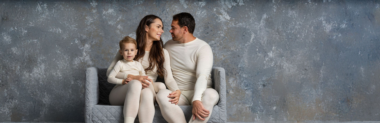 Family of three sitting together on a gray chair against a textured gray wall.