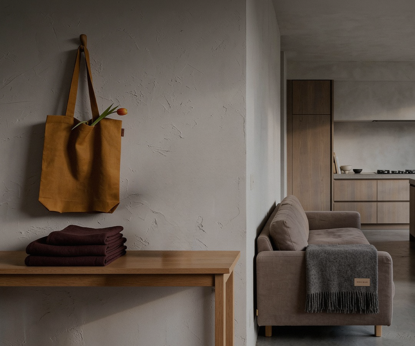 A vertical view of a minimalist, wabi-sabi style entryway leading into a living space. On the left, a textured off-white wall features a mustard yellow tote bag hanging from a wooden peg, with a single orange tulip peeking out. Below it is a light wood console table holding a stack of folded burgundy linens and a small ceramic vase with eucalyptus branches. To the right, the wall corners to reveal a partial view of a beige sofa draped with a grey fringed blanket, and a modern kitchen with warm wood cabinets