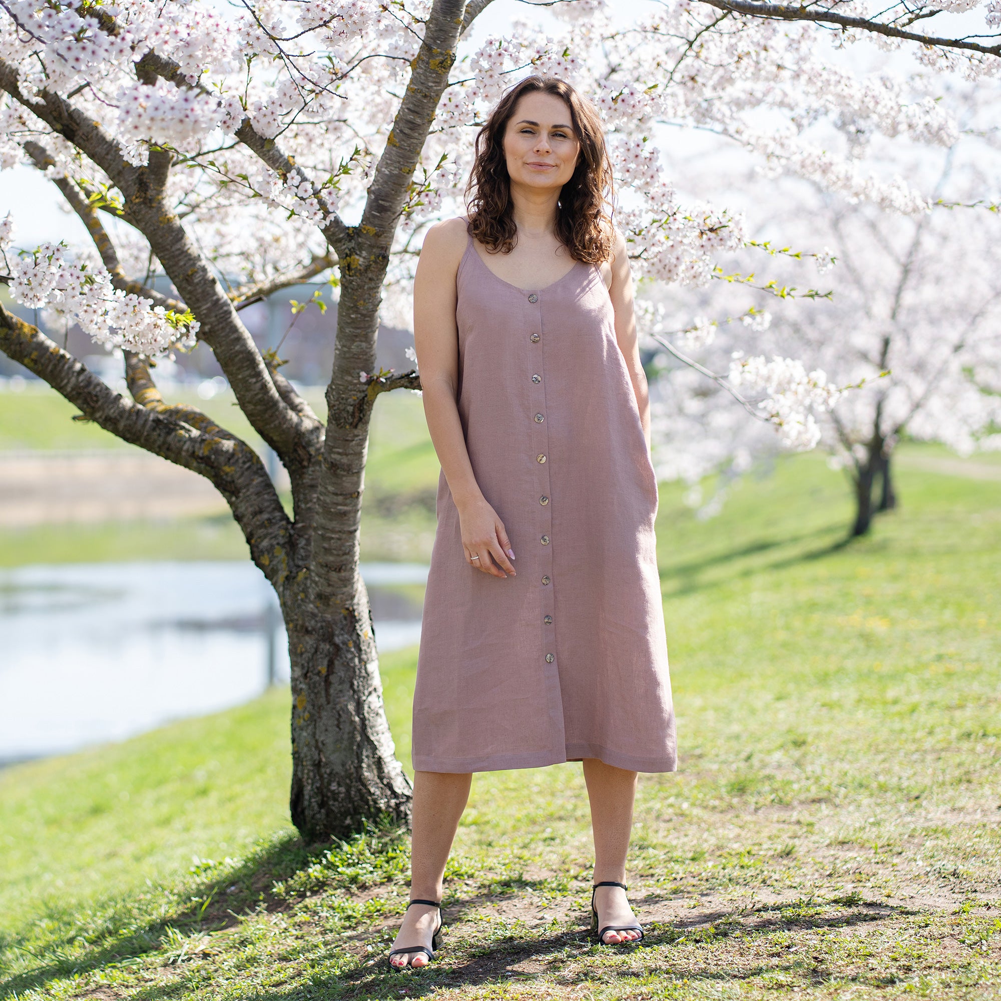 A woman with long, dark, wavy hair, wearing a sleeveless, button-front, faded rose midi-dress, stands outdoors in front of a tree with white blossoms. She holds a sprig of the blossoms up to her nose with her right hand. Her left hand is on her hip. She looks directly at the viewer with a slight smile. The background is a sunny, outdoor setting with green grass and more blossoming trees.