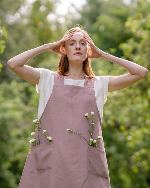 A fair-skinned person with reddish hair stands outdoors in a faded rose linen apron with white flowers in the pockets, hands touching their head, against blurred greenery.