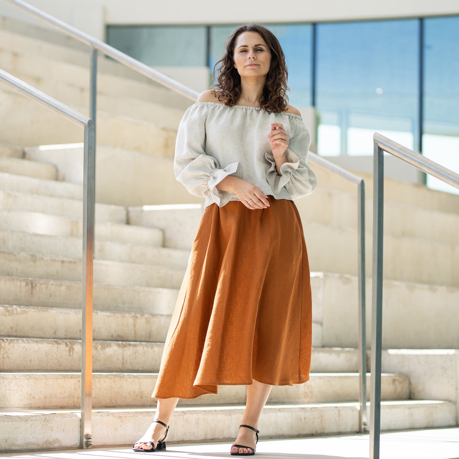 A woman with brown hair, wearing a light beige off-the-shoulder top with puff sleeves and a long rust-brown skirt, poses on concrete stairs.
