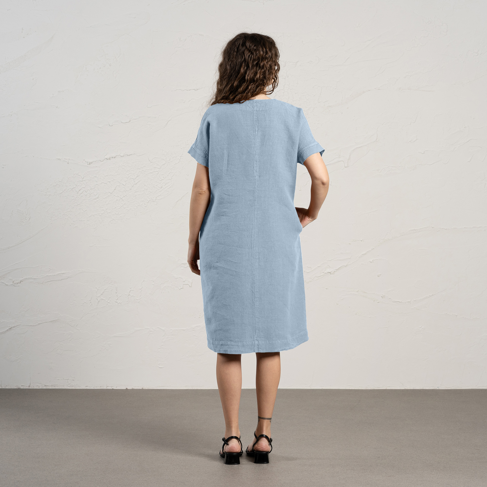 A woman with curly brown hair smiles while modeling a cloudy blue linen menique dress with short sleeves and pockets.