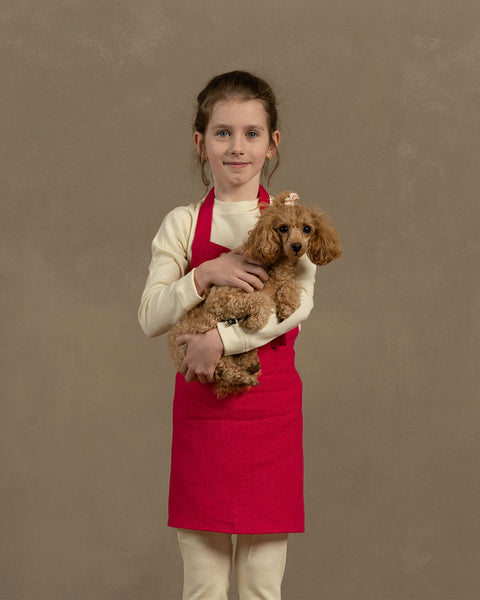 Young girl holding a small brown dog against a plain background wearing hot pink menique linen kids apron