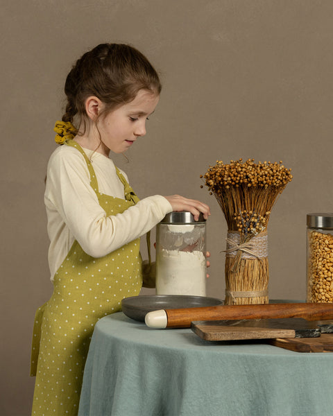 A child wearing a linen apron with leaf dots, standing at a table involved in crafting or baking, with ingredients and baking tools on the table.
