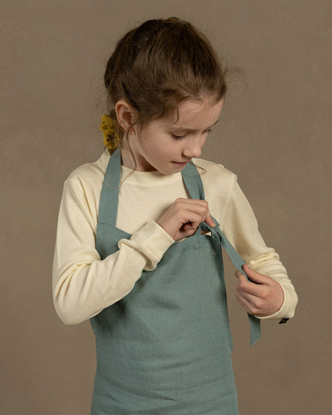 A young person with hair pulled back is shown from the waist up, looking down and adjusting the strap of a mint green apron. They are wearing a long-sleeved cream-colored shirt underneath. The background is a plain, light brown color.