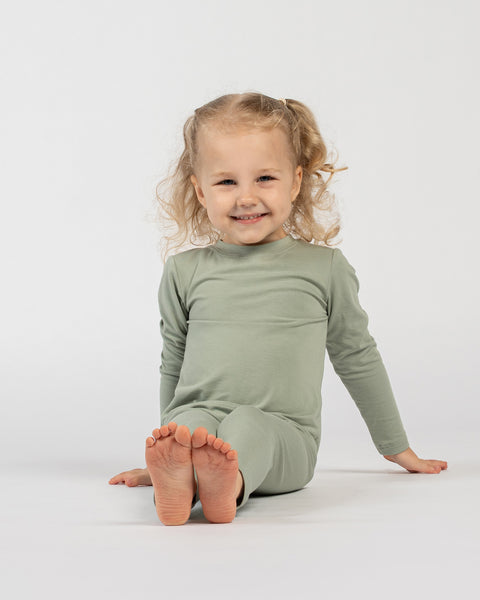 Smiling little girl wearing a green bamboo kids’ clothing set with long sleeves and pants, sitting barefoot on a white background.