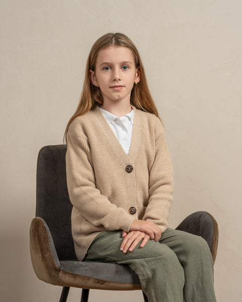 A girl is sitting on a gray chair, with her hands put neatly on her knees. She is wearing the menique kids&#39; merino wool cardigan in beige color. The picture emphasizes the v-neck shape of the cardigan.