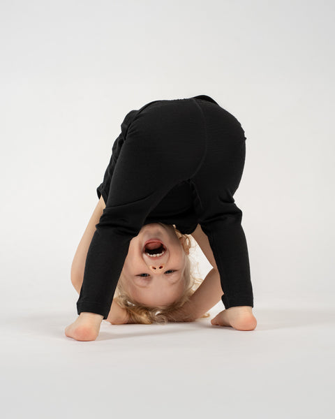 A young child with light-colored hair is playfully bent over, looking between their legs, against a plain white background. They are wearing dark-colored pants. The child&#39;s head is inverted, and they are smiling widely with their mouth open, looking directly at the camera. Their bare feet are visible on the floor.
