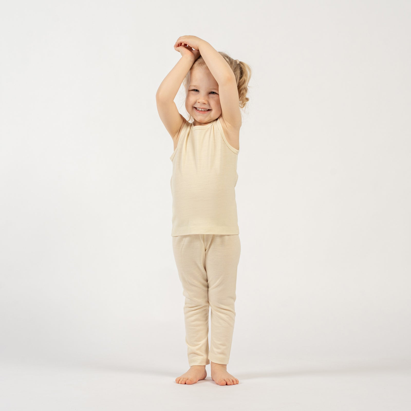A girl standing by a yellow armchair, holding a teddy bear by the hand, wearing the menique Kids' 160 Merino Pants in Natural color.