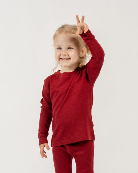 A young child, with light-colored hair pulled back, is shown from the waist up against a plain white background. The child is smiling broadly and looking towards the viewer. They are wearing a long-sleeved, menique merino wool top and matching red pants, suggesting pajamas or loungewear. 