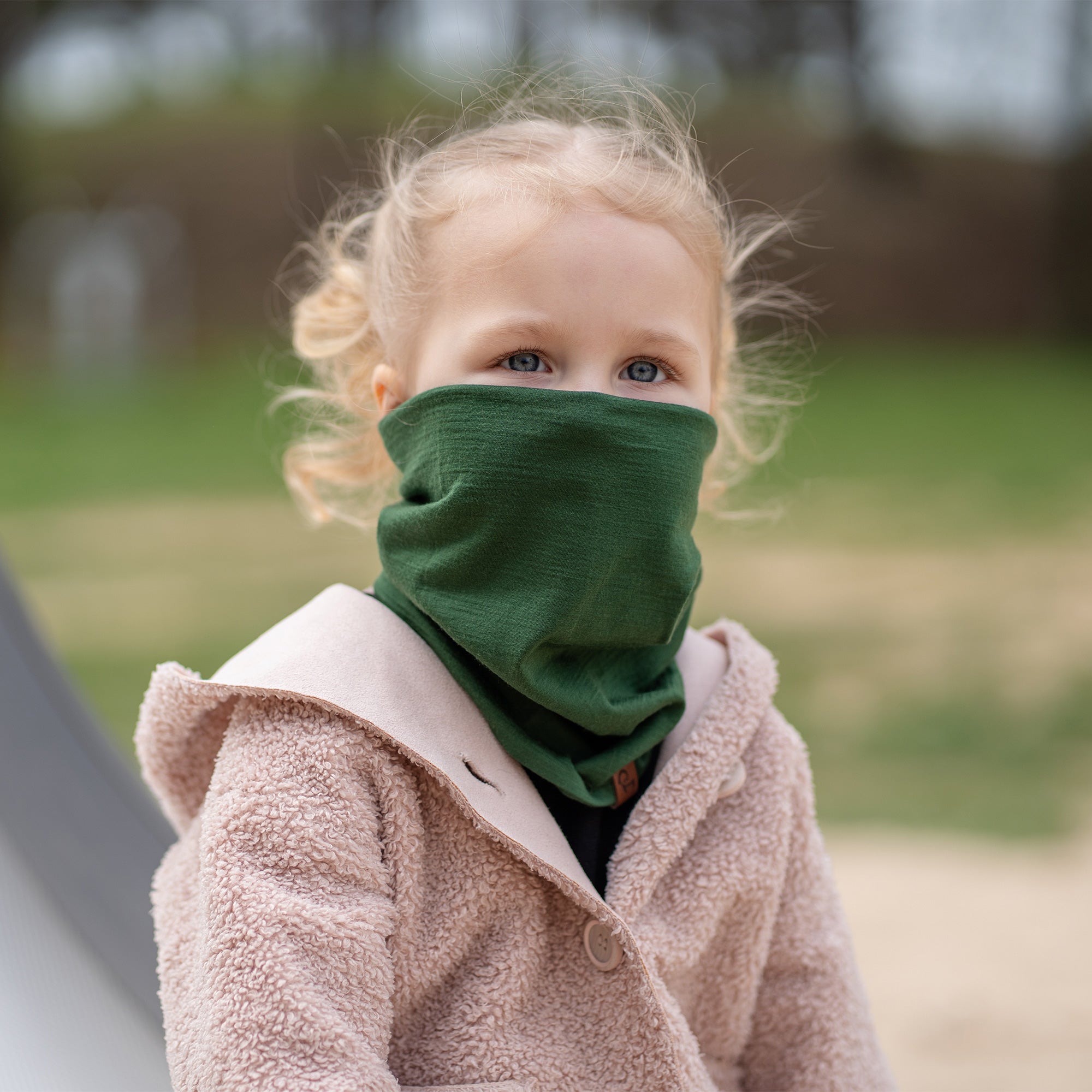 A young child with blonde hair tied in pigtails looks upwards and to the side, wearing a dark green Menique neck gaiter pulled up over their nose and mouth, paired with a beige hooded jacket. The child's blue eyes are visible above the neck gaiter, suggesting a moment of curiosity or observation outdoors.