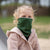 A young child with blonde hair tied in pigtails looks upwards and to the side, wearing a dark green Menique neck gaiter pulled up over their nose and mouth, paired with a beige hooded jacket. The child's blue eyes are visible above the neck gaiter, suggesting a moment of curiosity or observation outdoors.