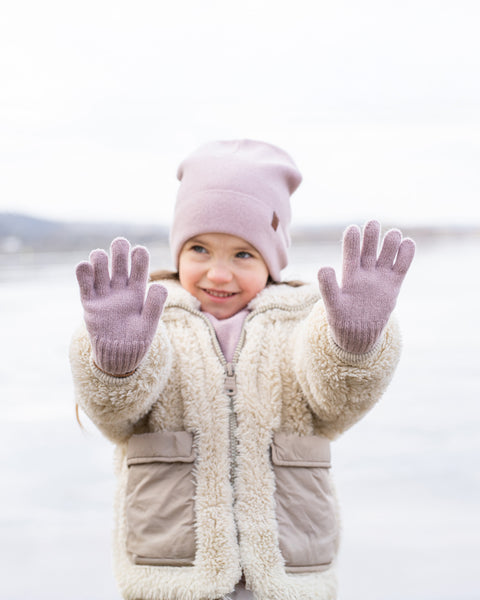 A young child with a joyful expression and slightly upward gaze, stands outdoors with both hands raised, palms facing forward. They are wearing a light pink knit beanie, a cream-colored sherpa coat with beige pockets, and light pink knitted gloves. The background is a blurred, bright, wintry landscape.